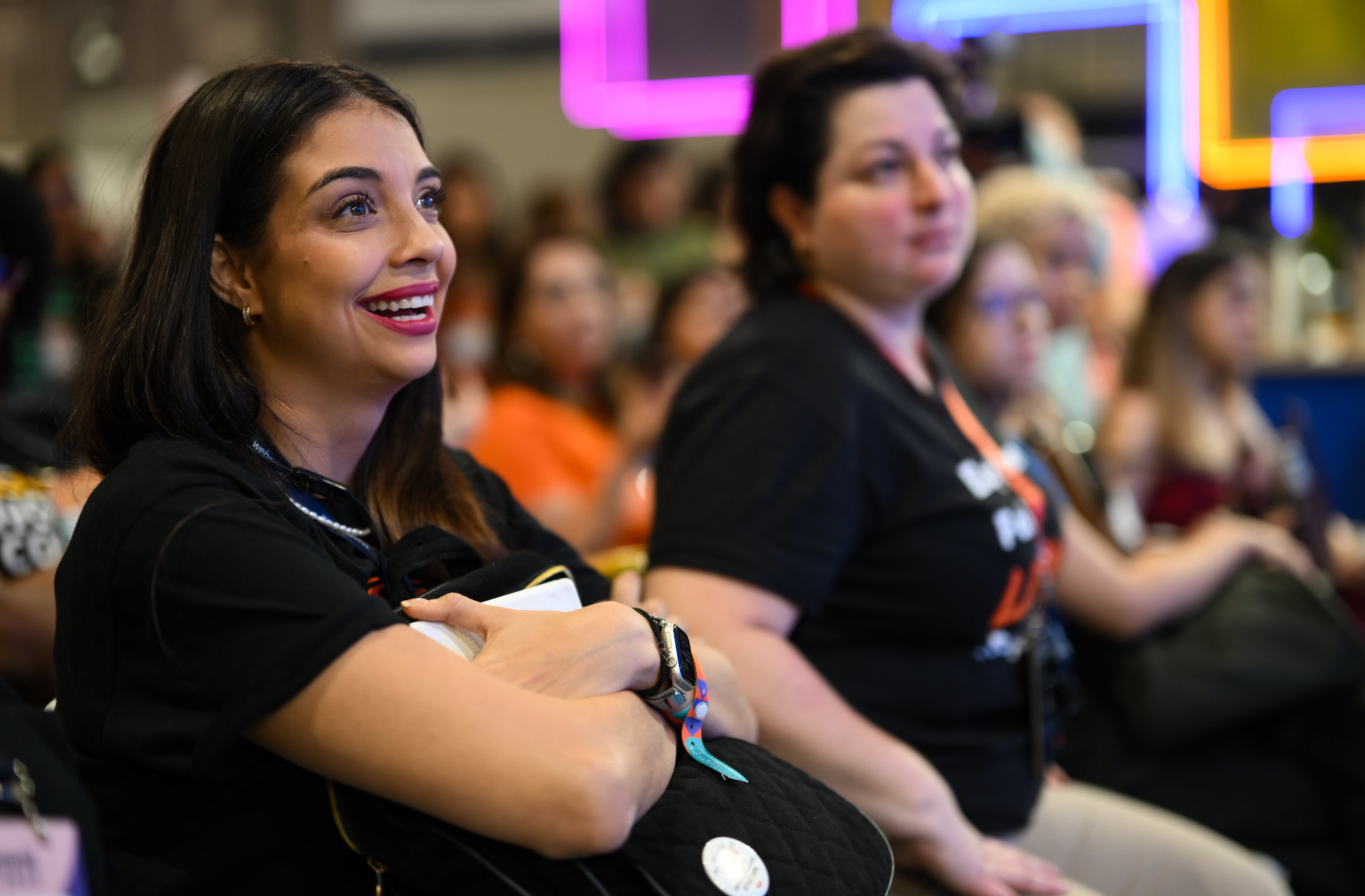 A smiling person hugs a backpack and gazes at something out of frame. Beyond this person are a row of other people who appear to be sitting down. This is the audience at a stage in the Web Summit Rio Women in Tech Lounge.