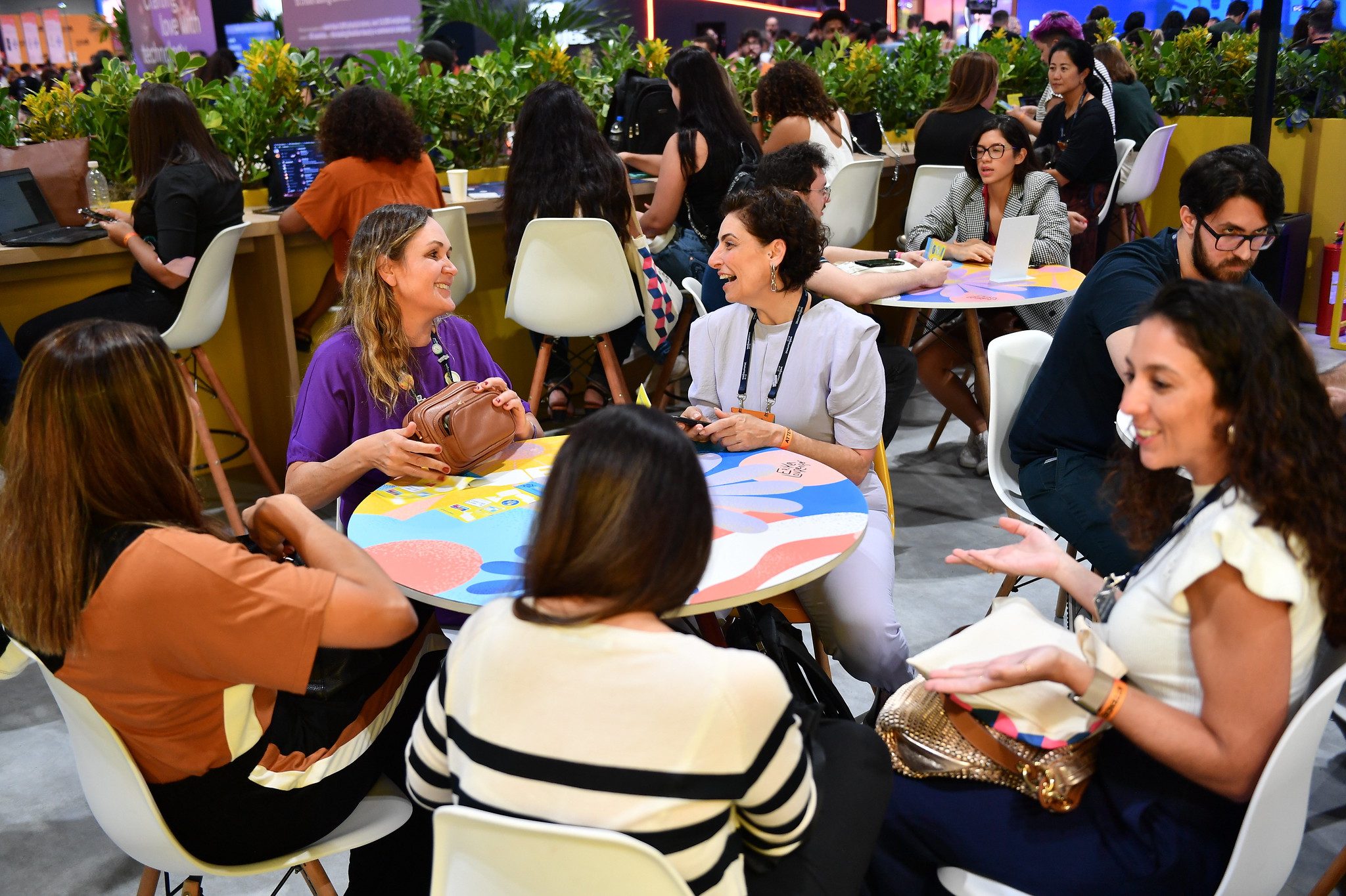 A group of smiling and laughing people sit around a round table. They appear to be engaged in several conversations. In the space around them are people sitting at other tables, and a row of people sitting on high stools at a counter. This is the Women in Tech Lounge at Web Summit Rio 2023.