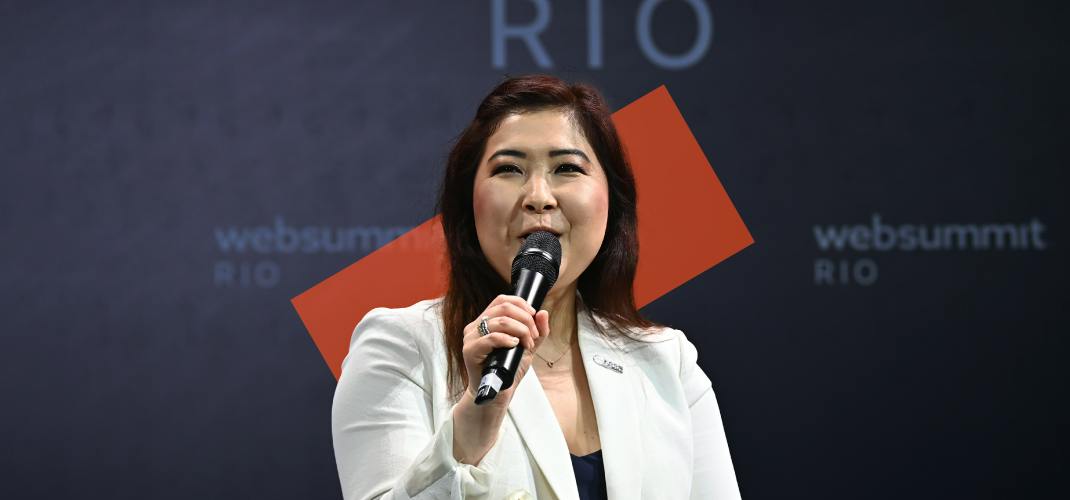 An image of Sandra Ro, CEO of Global Blockchain Business Council. Sandra is holding a microphone in one hand and appears to be speaking. The Web Summit Rio logo appears in several places on a wall behind Sandra.