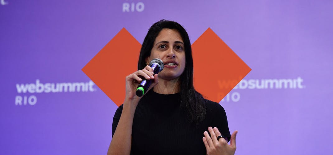 A person (Izabel Gallera, partner at Canary) is holding a microphone and appears to be talking. There is a purple background with white text (Web Summit Rio) and a semi transparent cross shaped design superimposed in the background.