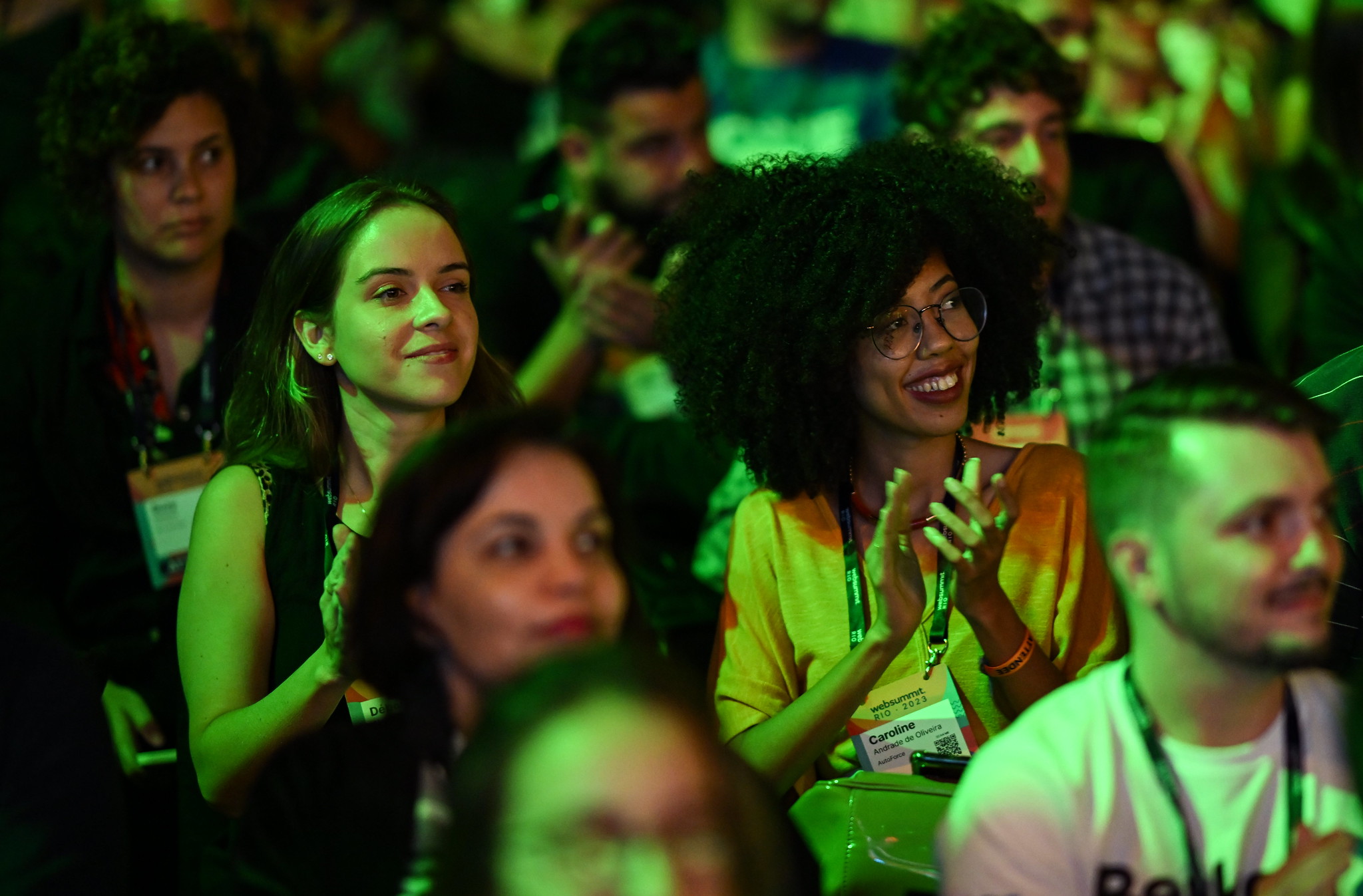 Two people sitting in a crowded audience. Both are smiling and clapping. They – and the rest of the audience – are looking at something out of frame. This is Web Summit Rio.