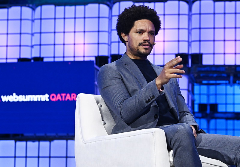 A person (author and presenter Trevor Noah) sitting in an armchair. Trevor is wearing a headset mic and gesturing with his right hand. Behind Trevor, a screen reads 'Web Summit Qatar'. This is Opening Night of Web Summit Qatar.