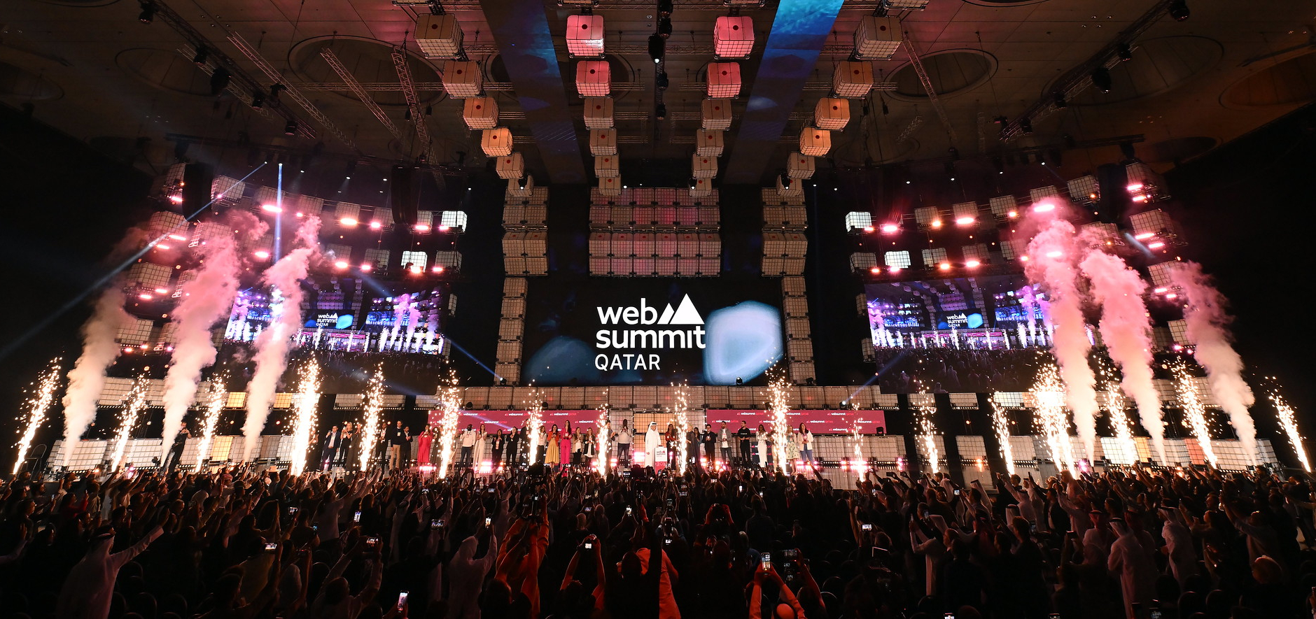 A large stage viewed over the heads of a crowded audience who are on their feet. On the stage, a long line of people face the audience. At the front of the stage, pyrotechnics go off. A large screen hanging at the back of the stage shows the Web Summit Qatar logo. This is Opening Night of Web Summit Qatar.