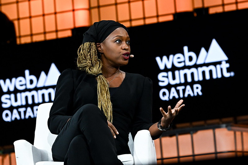A person (Africa Communications Media Group co-founder and CEO Mimi Kalinda) sitting in an armchair. Mimi is wearing a headset mic and gesturing with her left hand. She appears to be speaking. Screens behind Mimi bear the Web Summit Qatar logo. This is Centre Stage on Day 3 of Web Summit Qatar.