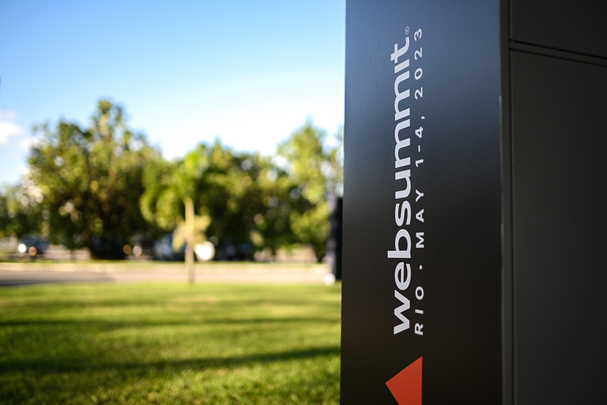 Photograph of Web Summit branding on a black object outside. To the left, there are blurred trees and a green area.