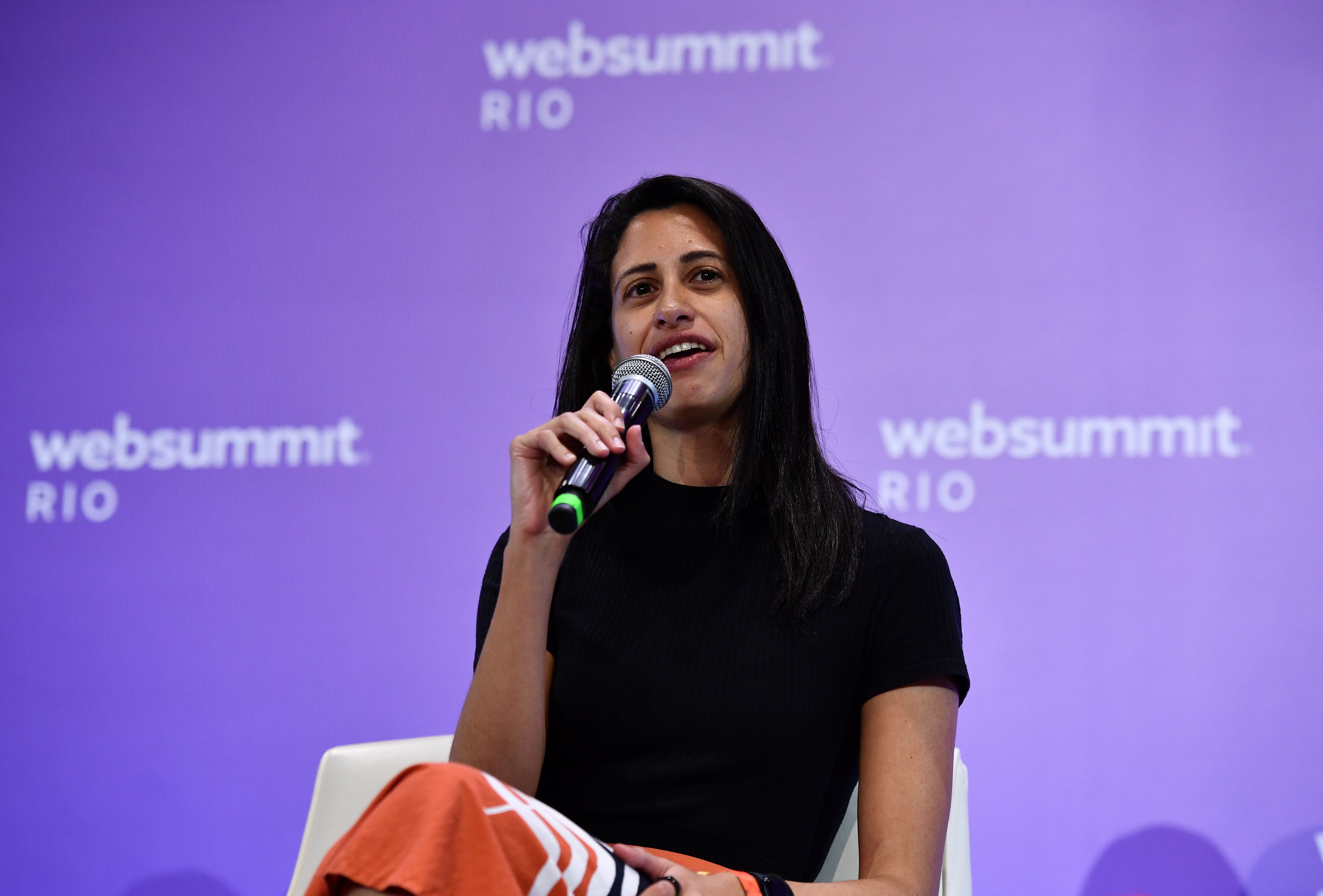 A person (Canary partner Izabel Gallera) sitting in an armchair on a stage. They are holding a microphone in their right hand, and appear to be speaking. The Web Summit Rio logo is visible in several places on the wall behind them.