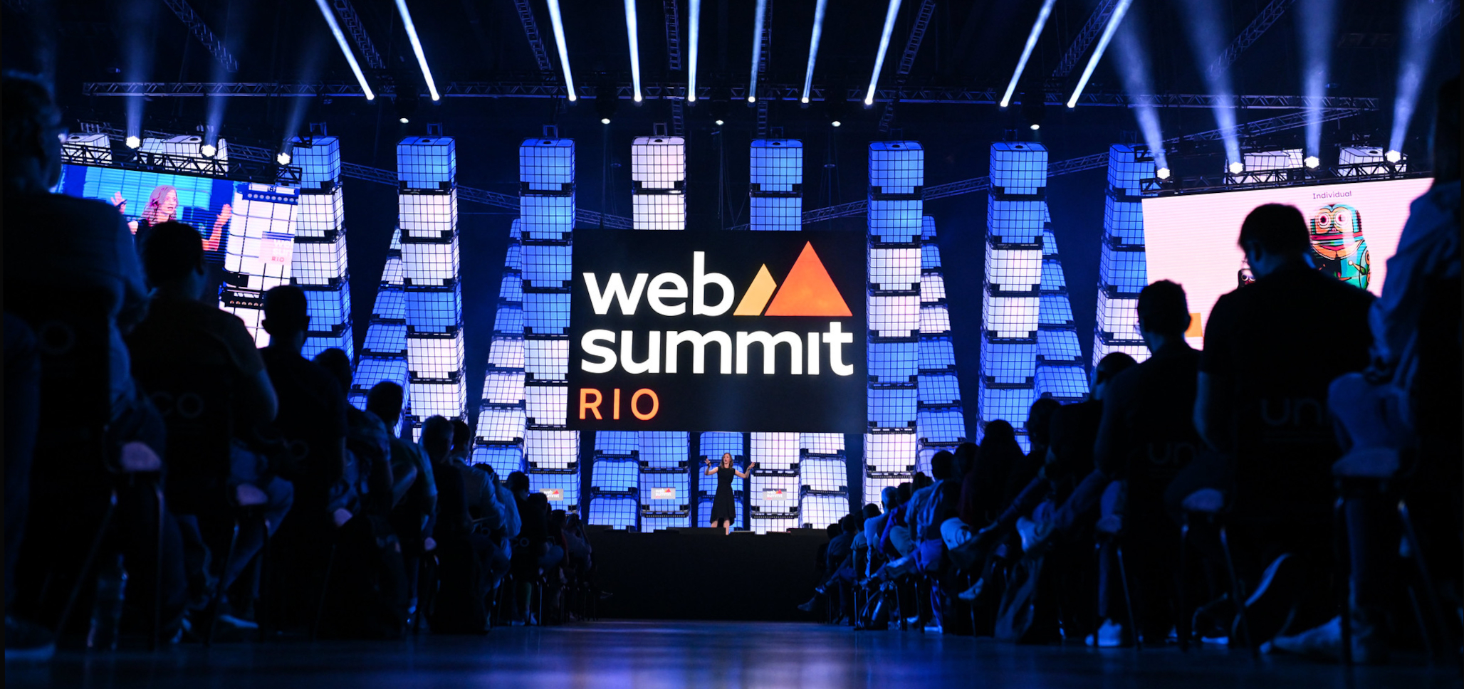 A person stands on a big stage, under a large Web Summit Rio sign. The stage is viewed from an aisle that runs through a crowded audience, which is seated.