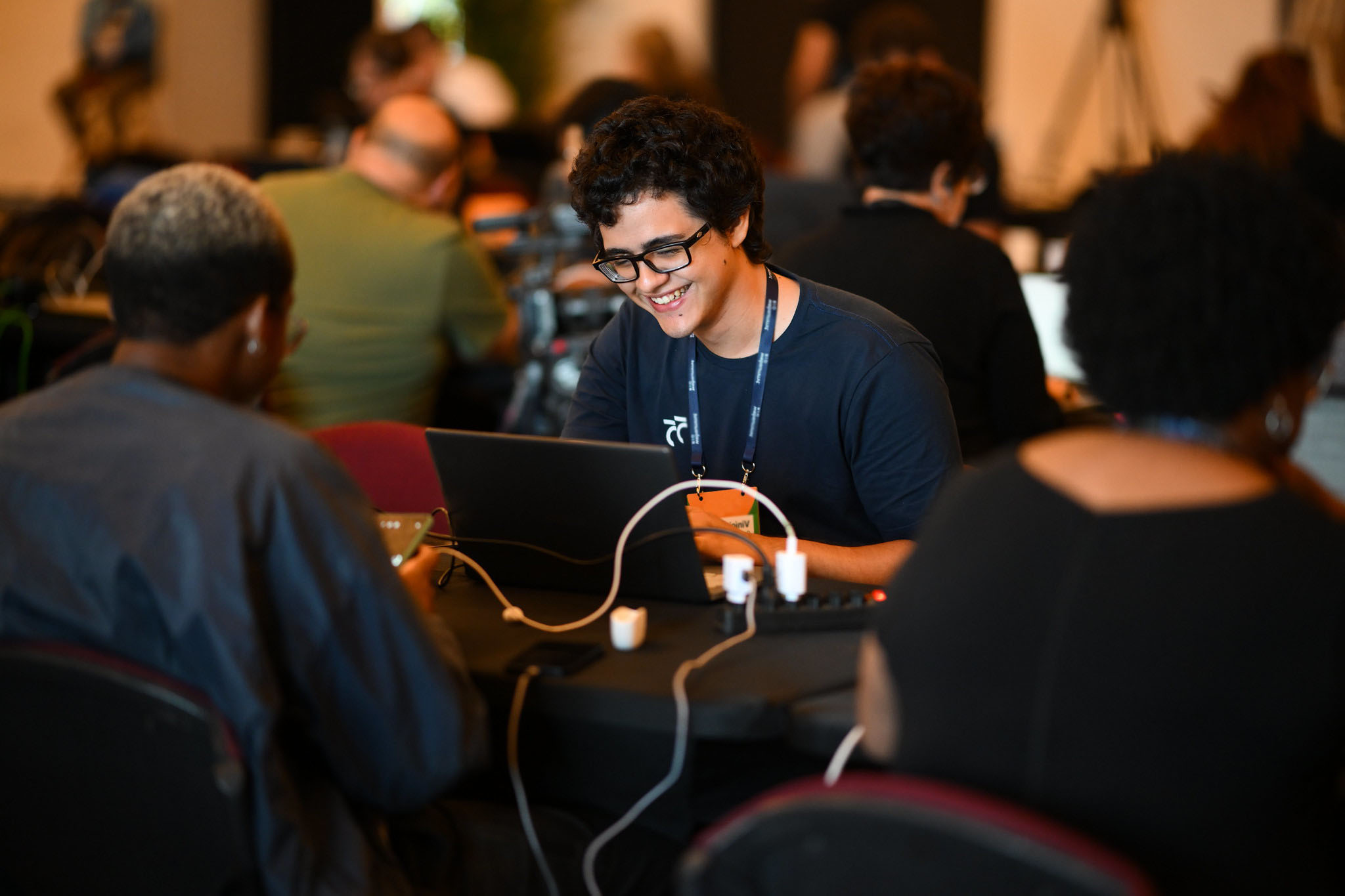 A smiling person working on a laptop in a room full of seated people.