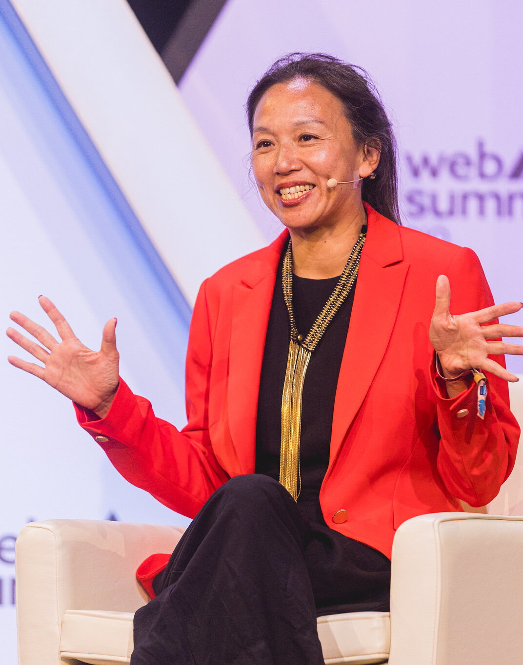 Photograph of a person (Bedy Yang of 500 Global) sitting on a chair onstage at Web Summit Rio. The person appears to be speaking and is gesturing with their hands. They are wearing a headset microphone.