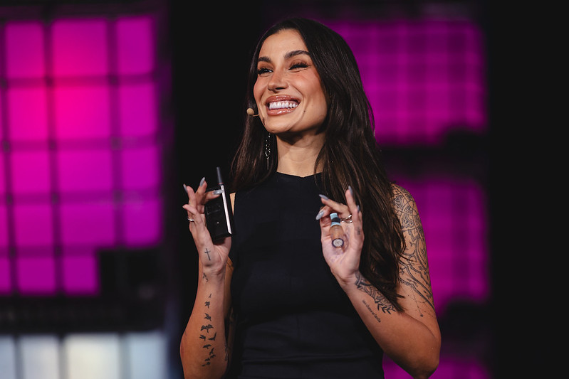 A smiling person (influencer Bianca Andrade) stands on stage. They are wearing a headset mic. They hold a presentation clicker in their right hand and what appears to be a tube of lipgloss in their left. They appear to be speaking. This is Center Stage at Web Summit Rio.