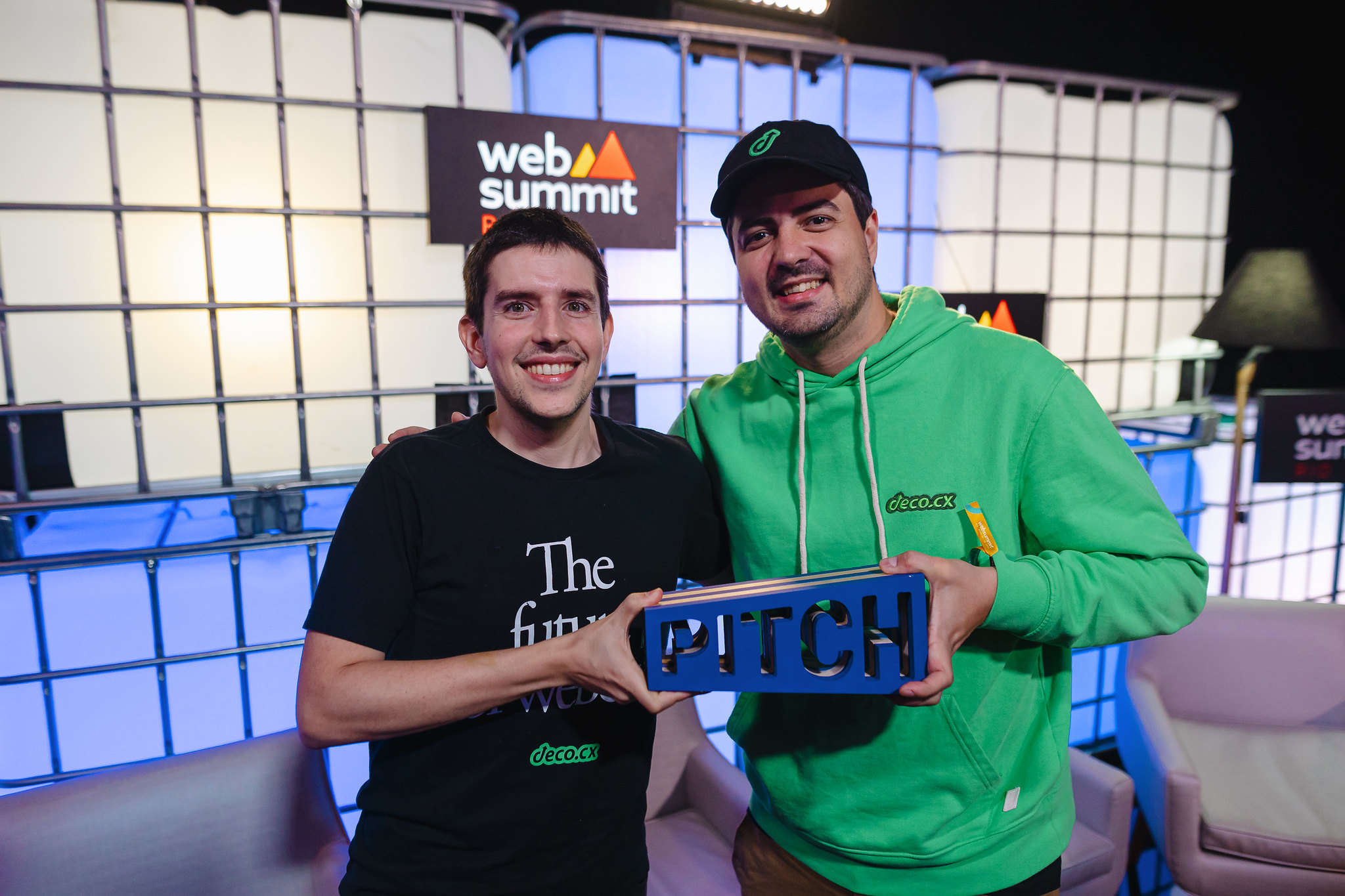 Two people (deco.cx co-founders Guilherme Rodrigues and Rafael Crespo) stand shoulder to shoulder. They are holding a wooden trapezoidal trophy with the word PITCH carved through it. Behind them is a wall of large cuboid water containers lit from within. deco.cx was the PITCH winner at Web Summit Rio 2024. This is backstage at Web Summit Rio's Center Stage.