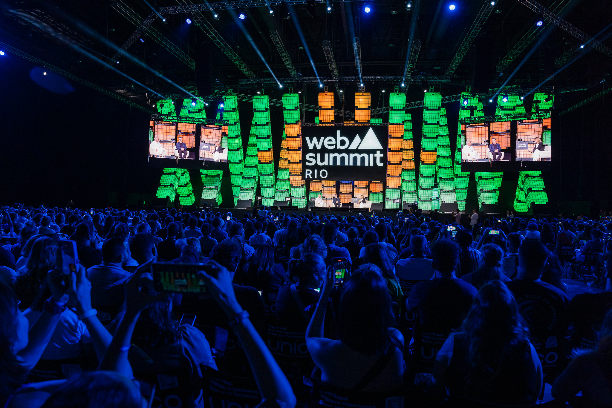 Image of Center Stage from Web Summit Rio 2024. The stage is brightly lit in green and orange. There are figures on stage in the distance. In the foreground, audience members can be seen standing, watching and holding phones aloft to take photos.