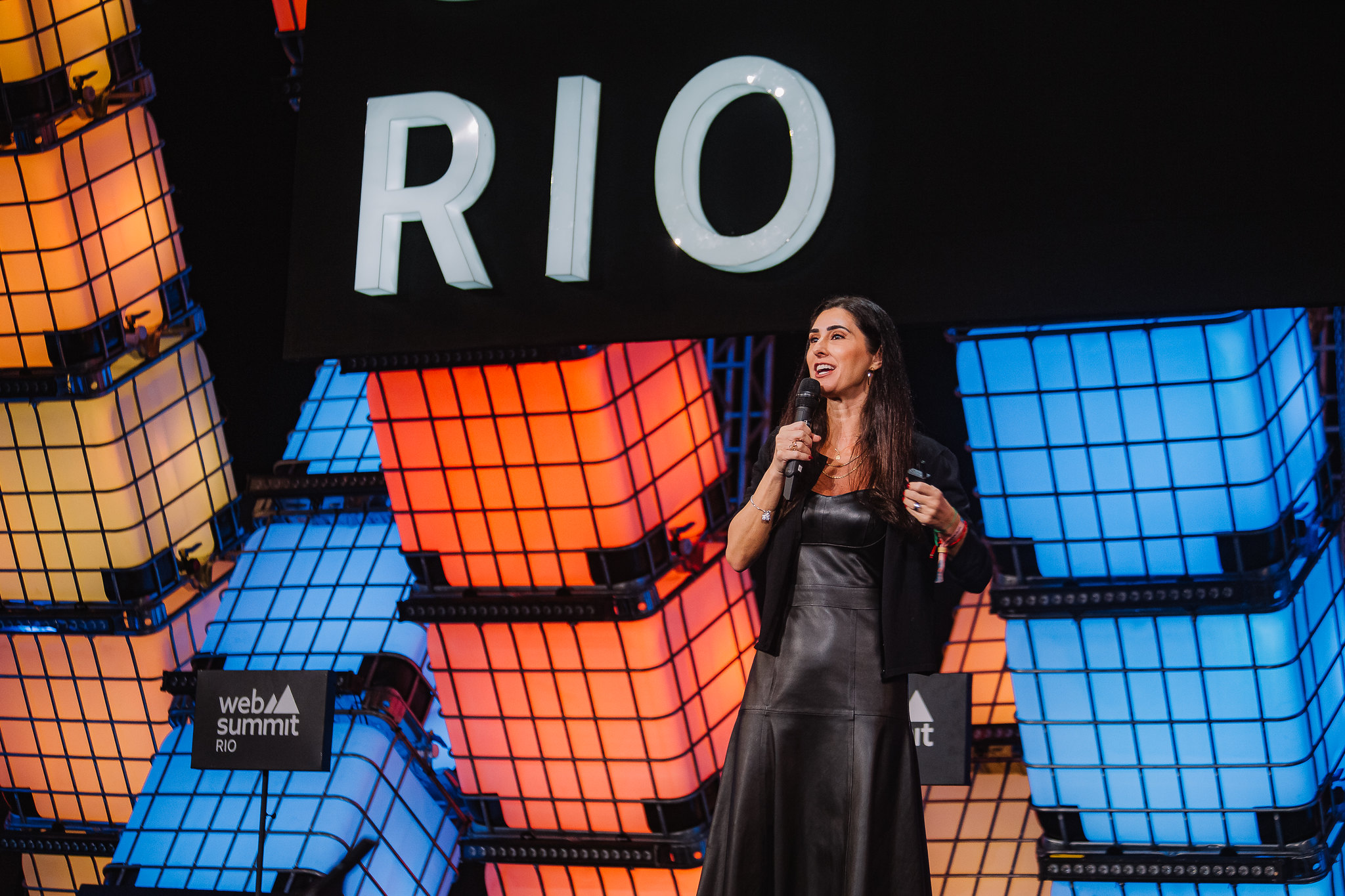 A person (TikTok general manager of global business solutions for LatAm Gabriela Comazzetto) standing on a stage. They are holding a microphone in their right hand, and a presentation clicker in their left. Behind them is a wall of large cuboid water containers, lit from within. Over the head is visible the word 'Rio', part of the Web Summit Rio logo. This is Center Stage at Web Summit Rio.