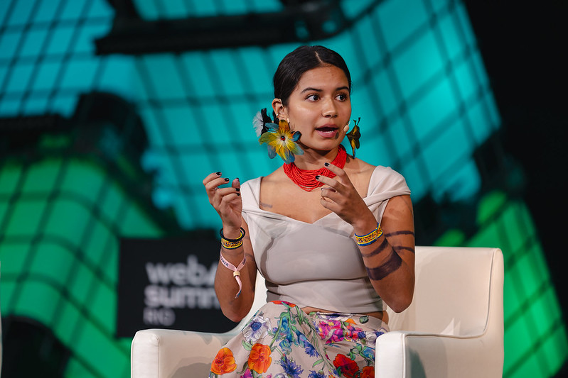 A person (Indigenous Youth Collective of Amazon Defenders co-founder Helena Gualinga) sits in an armchair. They are wearing a headset mic, and gesturing with both hands. They appear to be speaking. Behind them is a wall of large cuboid water containers, lit from within. This is Center Stage at Web Summit Rio.